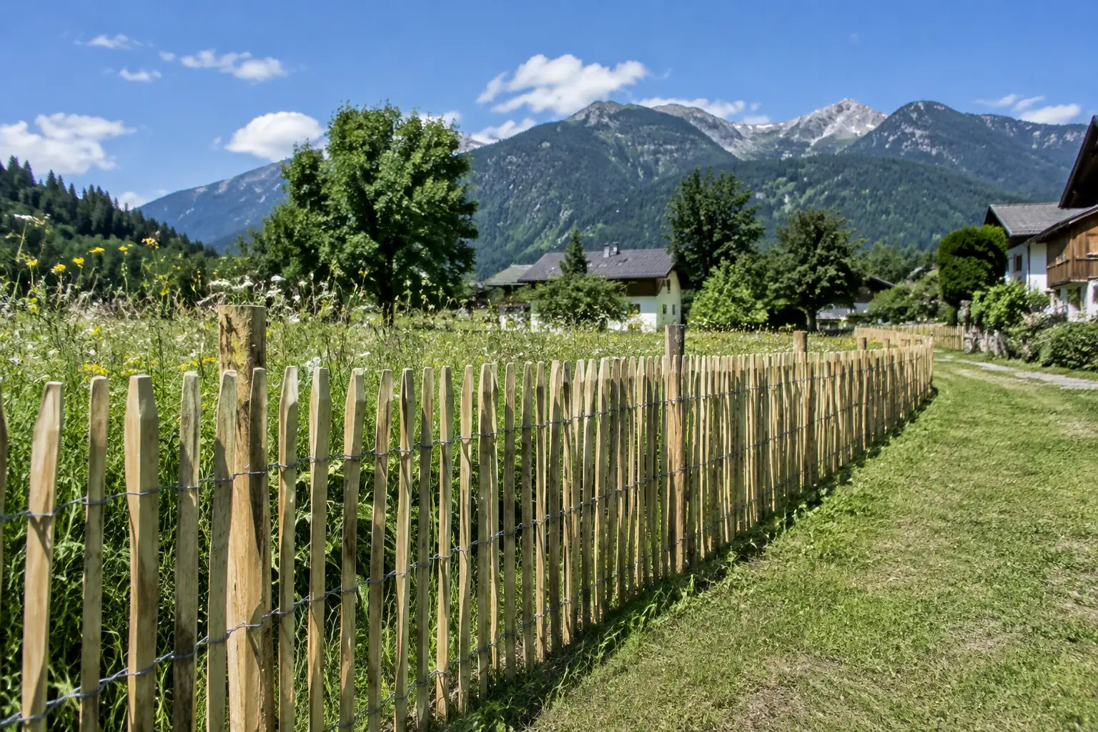 Clôture neuve en ganivelle de châtaignier liée au fil de fer, le long d’un pré en bordure de village, montagnes boisées au fond sous un ciel bleu