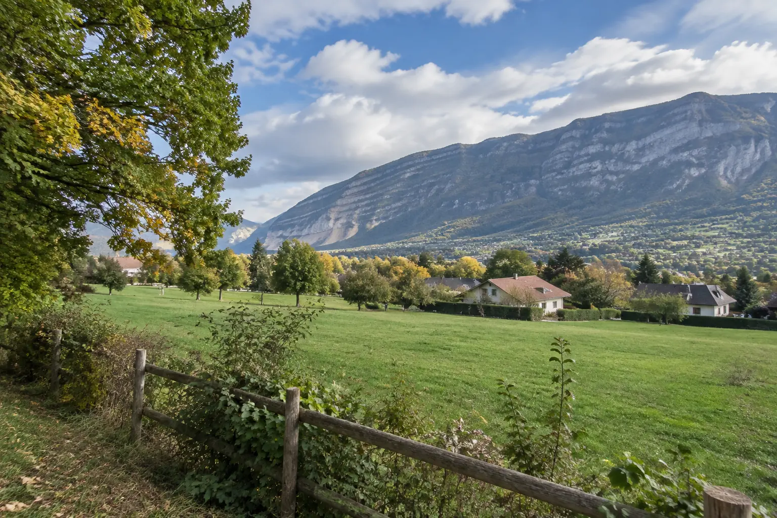 Lisière de Saint-Julien-en-Genevois : haie bocagère de chênes et d’érables champêtres le long d’un pré, clôture en bois, maisons dispersées et longue crête du Salève derrière, début d’automne