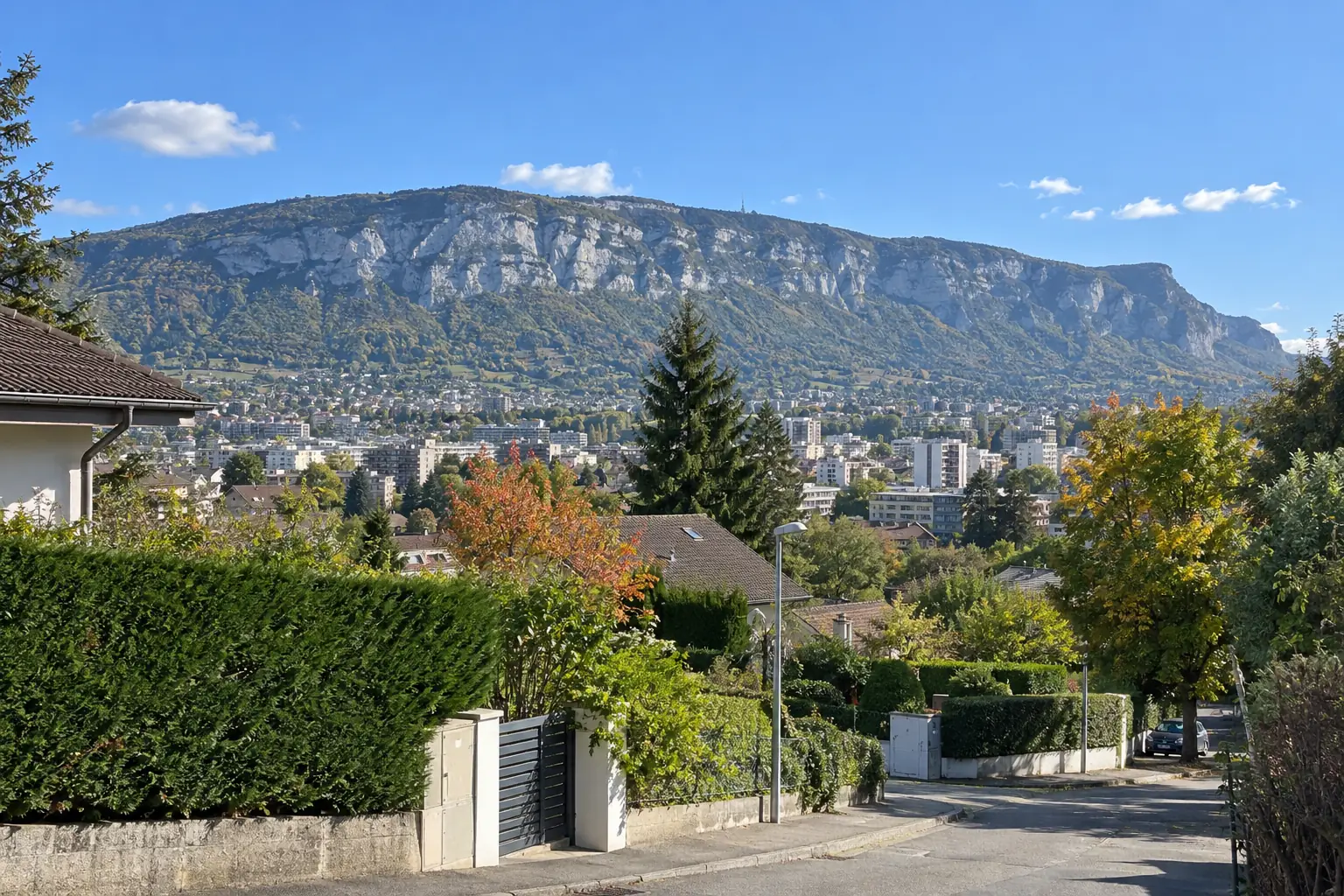 Vue depuis une rue résidentielle des hauteurs d’Annemasse : haies, toits et immeubles de la ville, falaises calcaires du Salève sous un ciel bleu